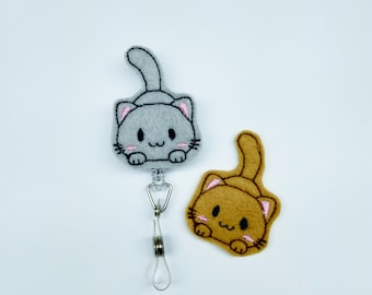 Peut inclure: Deux porte-clés en feutre en forme de chat, l'un gris et l'autre marron, avec un porte-badge rétractable argenté. Les chats sont brodés avec des yeux noirs et des joues roses.