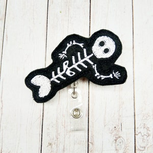 May include: Black felt skeleton with white embroidery, attached to a clear plastic retractable badge holder.