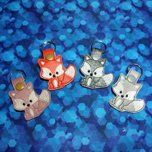 May include: Four embroidered fox keychains in shades of red, gray, and pink. Each keychain features a fox design with a white belly and tail, attached to a leather strap and metal ring. The background is a blue geometric pattern.