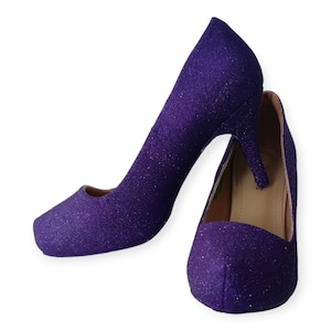 Puede incluir: Un par de zapatos de tac&oacute;n alto de purpurina morada. Los zapatos tienen punta puntiaguda y tac&oacute;n de aguja.