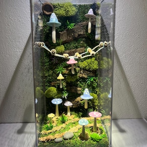 May include: A glass terrarium with a miniature forest scene featuring moss, small trees, and colorful mushroom figurines. The terrarium is decorated with a string of lights and small wooden beads.