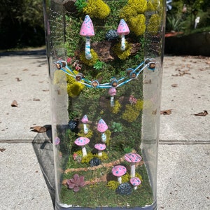 May include: A clear glass terrarium with a miniature forest scene. The terrarium features pink and blue mushrooms, green moss, and a small succulent plant.