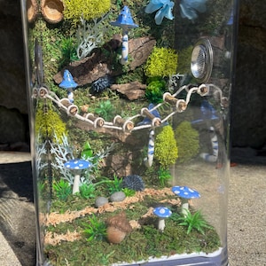May include: A miniature glass terrarium with a blue and white mushroom theme. The terrarium is filled with moss, small logs, and a rope bridge. There are several blue and white mushrooms with white polka dots scattered throughout the terrarium.