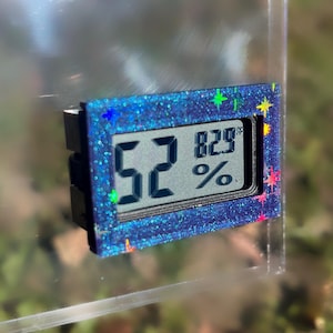 May include: A small digital hygrometer with a blue glitter frame displays 52% and 82.9&deg;F. The device has a black backing and colorful star-shaped accents. The screen shows the humidity and temperature readings.