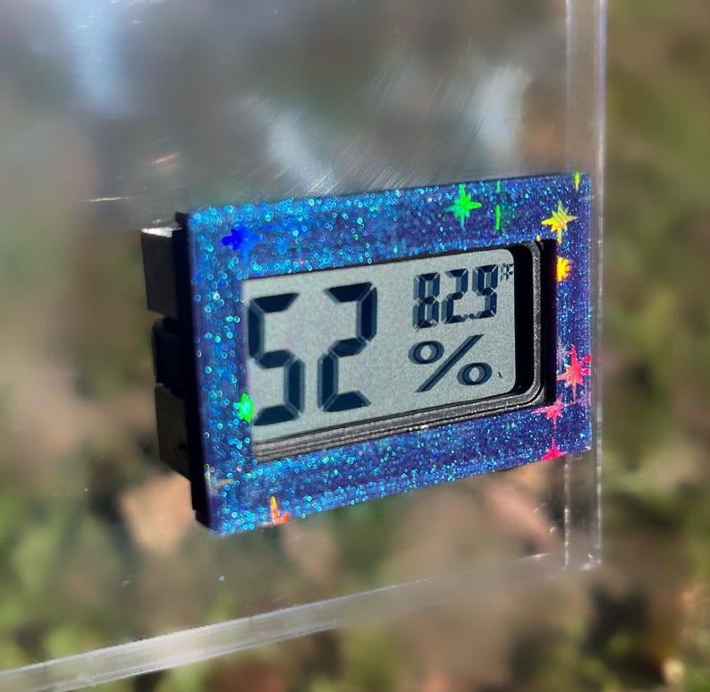 May include: A small digital hygrometer with a blue glitter frame displays 52% and 82.9&deg;F. The device has a black backing and colorful star-shaped accents. The screen shows the humidity and temperature readings.