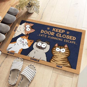 May include: A rectangular doormat with a navy blue background and a brown border. The mat features cartoon cats and the text "KEEP DOOR CLOSED...CATS PLANNING ESCAPE." The cats are named Oliver, Buddy, Charlie, and Max.