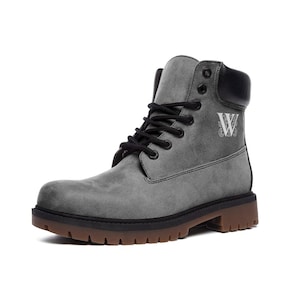 Könnte beinhalten: Graue Wildlederstiefel mit schwarzen Schnürsenkeln und einem schwarzen Ledermanschetten. Die Stiefel haben eine braune Gummisohle und ein weißes Monogramm "W" an der Seite.