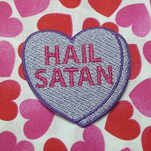 May include: A heart-shaped patch with the text "HAIL SATAN" embroidered in pink thread. The patch is gray with a purple border and is set against a background of red and pink hearts.