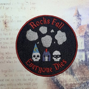 May include: Black, round embroidered patch with the text "Rocks Fall Everyone Dies" in red. The patch features three skulls, one with a wizard hat, one with a helmet, and one with a mask, along with gray rock designs.
