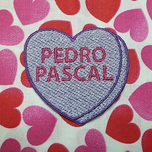 May include: A purple heart-shaped candy with the text "PEDRO PASCAL" in pink lettering on a white background with a pink and red heart pattern.