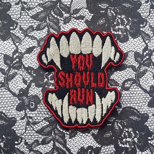 May include: A black and white embroidered patch with a red outline. The patch features a set of teeth and the text "You Should Run".