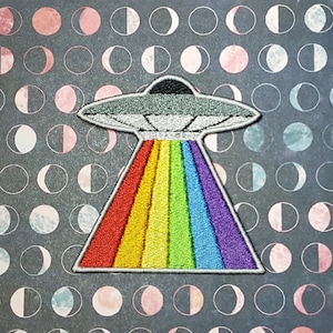May include: An embroidered patch featuring a gray UFO saucer shape at the top with a wide, triangular beam of light extending downward. The beam is composed of vertical stripes in red, orange, yellow, green, blue, and purple rainbow colors.