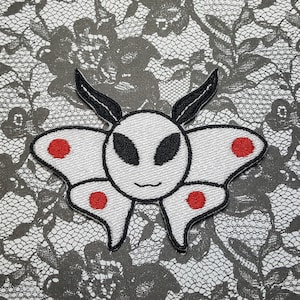 May include: Black and white embroidered patch of a moth with red dots and antennae. The moth has a white face with a smiling mouth.