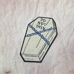 May include: A black and white embroidered patch of a closed coffin with the text "Do Not Open" on the top. The coffin is tied with a blue chain.