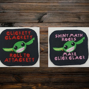 May include: Two embroidered patches with a cartoon character. One patch has the text "CLICKETY CLACKETY ROLL TO ATTACKETY" and the other says "SHINY MATH ROCKS MAKE CLICK CLACK". The character is green with large eyes.