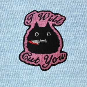 May include: Pink and black embroidered patch featuring a black cat holding a knife in its mouth. The text "I Will Cut You" is written around the cat.