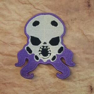 May include: Purple and white embroidered patch of a skull with tentacles. The skull has a toothy grin and black eyes.