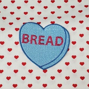 Può includere: Un cuore di caramella blu con la parola "BREAD" ricamata in rosa su uno sfondo bianco con un motivo a cuore rosso.