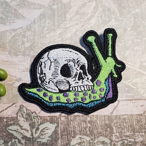 May include: A green and purple embroidered iron-on patch of a snail with a skull for a shell.