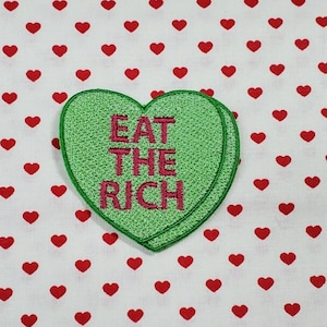 Puede incluir: Parche verde en forma de corazón con el texto "EAT THE RICH" en letras rosas.