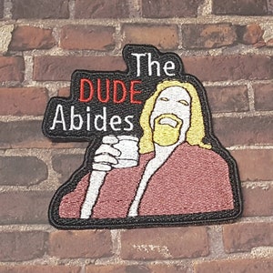 May include: Embroidered patch featuring a cartoon of The Dude from The Big Lebowski, holding a glass of White Russian. The text "The Dude Abides" is written in red and white on a black background.