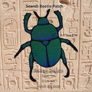 Puede incluir: Un parche bordado de escarabajo con hilo azul y verde y contornos negros. El texto dice: Scarab Beetle Patch, 8,89 cm x 7,62 cm, Backing Options, Sew-on, Iron-on, Hook & Loop. El parche se muestra sobre un fondo de pared de piedra.