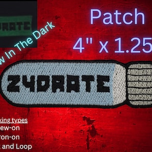 Könnte beinhalten: Ein weißer und grauer gestickter Patch mit dem Wort "ZYDRATE" in schwarzen Buchstaben. Der Patch hat die Form einer Pille und verfügt über eine Leucht-im-Dunkeln-Funktion. Der Patch ist 10,16 cm x 3,18 cm groß und in den Ausführungen zum Annähen, Bügeln und mit Klettverschluss erhältlich.