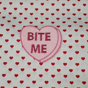 Puede incluir: Parche rosa en forma de corazón con el texto "BITE ME" en bordado rosa sobre un fondo blanco con un patrón de corazones rojos.