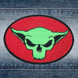 Könnte beinhalten: Grüner Schädel mit Hörnern, auf einem roten Kreis-Patch gestickt. Der Patch ist auf einen blauen Jeansstoff genäht.