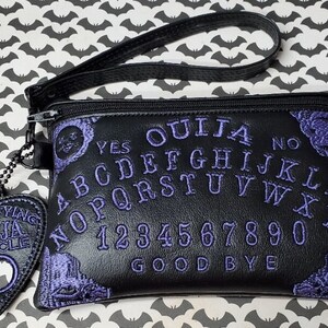 Puede incluir: Bolsa de cuero negro con cremallera con un dise&ntilde;o de tablero de Ouija bordado en morado. La bolsa tiene una correa negra y un amuleto en forma de coraz&oacute;n morado con las palabras "Mystifying Ouija Oracle".