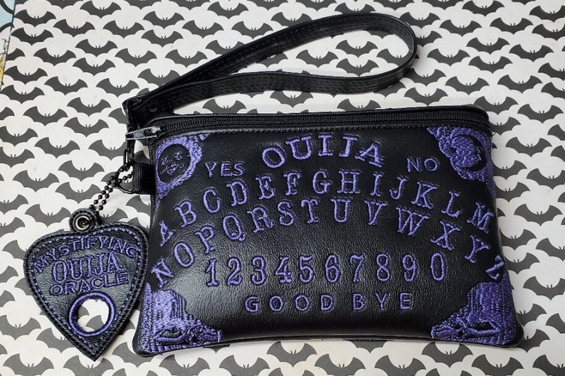 Puede incluir: Bolsa de cuero negro con cremallera con un dise&ntilde;o de tablero de Ouija bordado en morado. La bolsa tiene una correa negra y un amuleto en forma de coraz&oacute;n morado con las palabras "Mystifying Ouija Oracle".
