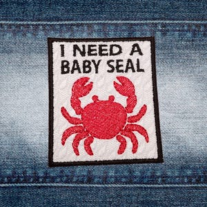 May include: Embroidered patch featuring the text "I NEED A BABY SEAL" above a red crab illustration. The patch has a white, textured background and a black border. It is set against a denim background.