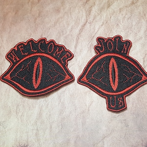 Puede incluir: Dos parches bordados en negro y rojo con un diseño de ojo. El primer parche dice "WELCOME" y el segundo parche dice "JOIN US".