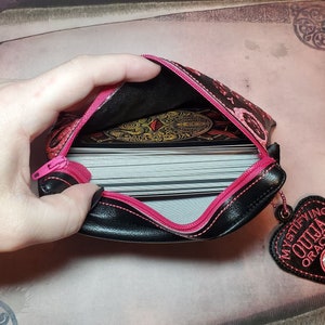 Puede incluir: Una bolsa negra con cremallera con una cremallera rosa y ribete rosa. La bolsa est&aacute; decorada con un patr&oacute;n floral y tiene un amuleto rosa en forma de coraz&oacute;n que dice "Mystifying Ouija Oracle". La bolsa est&aacute; abierta y hay una baraja de cartas dentro.