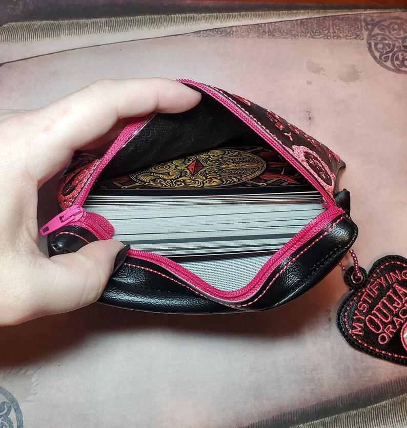 Puede incluir: Una bolsa negra con cremallera con una cremallera rosa y ribete rosa. La bolsa est&aacute; decorada con un patr&oacute;n floral y tiene un amuleto rosa en forma de coraz&oacute;n que dice "Mystifying Ouija Oracle". La bolsa est&aacute; abierta y hay una baraja de cartas dentro.