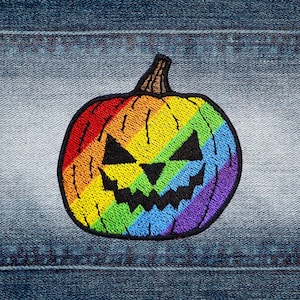 Op de afbeelding: Een geborduurde patch van een pompoen met een regenboogkleurig verloopmotief. De pompoen heeft een zwart geborduurd jack-o'-lantern gezicht met driehoekige ogen, een driehoekige neus en een gekartelde mond, omlijnd met zwart garen.