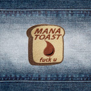 May include: A rectangular embroidered patch shaped like a slice of bread with a dark brown crust and a light tan center. The text MANA TOAST is embroidered in brown at the top, a brown teardrop shape in the center, and the text fuck u at the bottom.