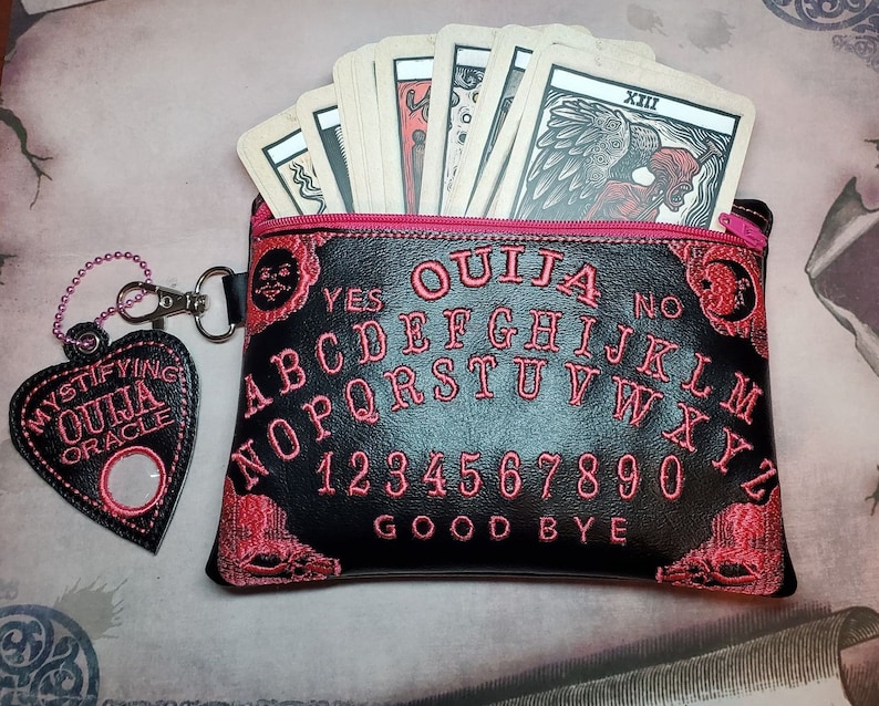Puede incluir: Una bolsa de cuero negro con una cremallera rosa y detalles bordados en rosa. La bolsa presenta un dise&ntilde;o de tablero Ouija en blanco y negro con las letras, los n&uacute;meros y las palabras "YES", "NO" y "GOODBYE". La bolsa est&aacute; abierta y contiene una baraja de cartas con una ilustraci&oacute;n en blanco y negro de una figura con alas.