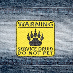 Può includere: Una toppa ricamata quadrata con sfondo giallo e bordo nero. La toppa presenta una grafica con impronta di zampa nera al centro, con il testo WARNING in alto e SERVICE DRUID DO NOT PET stampato in lettere nere nella parte inferiore.