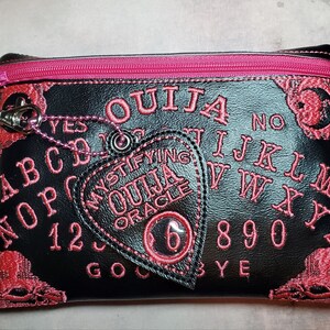 Puede incluir: Bolsa negra con cremallera con una cremallera rosa y un dise&ntilde;o de tablero de Ouija bordado en rosa. La bolsa incluye un amuleto rosa en forma de coraz&oacute;n con las palabras "Mystifying Ouija Oracle" y el n&uacute;mero "6" en su interior.