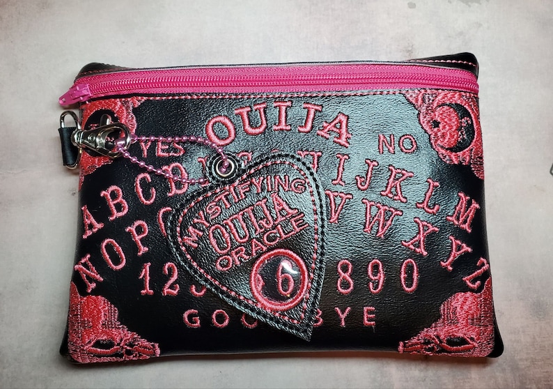 Puede incluir: Bolsa negra con cremallera con una cremallera rosa y un dise&ntilde;o de tablero de Ouija bordado en rosa. La bolsa incluye un amuleto rosa en forma de coraz&oacute;n con las palabras "Mystifying Ouija Oracle" y el n&uacute;mero "6" en su interior.