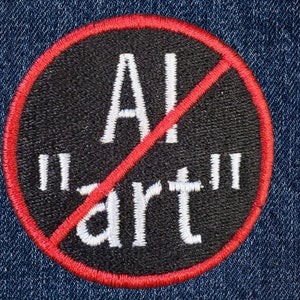 May include: A circular embroidered patch with a black background and a red border. The text "AI "art"" is stitched in white letters in the center. A diagonal red line crosses through the text, indicating a prohibition symbol over the words.