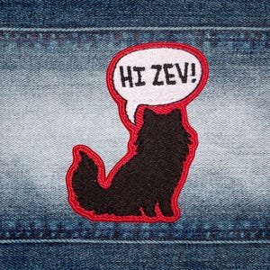 May include: Embroidered patch with a black cat silhouette and a red border. A white speech bubble above the cat reads "HI ZEV!" in black text. The patch is set against a denim background.