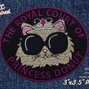 Puede incluir: Un parche bordado en negro y rosa con un gato de dibujos animados que lleva gafas de sol y una corona. El texto "THE ROYAL COURT OF PRINCESS DONUT" está escrito alrededor del gato. El parche mide 7,6 cm por 8,9 cm y se puede coser, planchar o sujetar con velcro.