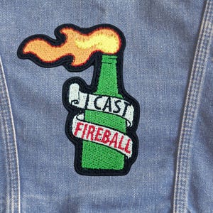 Può includere: Una bottiglia di vetro verde con una fiamma in cima e uno striscione bianco che dice "I Cast Fireball".