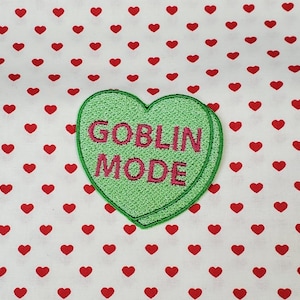 Può includere: Toppa a forma di cuore verde con testo ricamato rosa che recita "GOBLIN MODE" su uno sfondo bianco con cuori rossi.