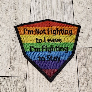 Puede incluir: Un parche bordado con un diseño de arcoíris y el texto "I'm Not Fighting to Leave I'm Fighting to Stay".