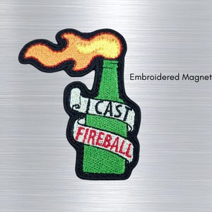 Könnte beinhalten: Ein bestickter Magnet mit einer grünen Flasche und einer gelben sowie orangefarbenen Flamme an der Spitze. Ein weißes Banner umwickelt die Flasche mit dem schwarzen Text CAST und dem roten Text FIREBALL. Der Text Embroidered Magnet! ist sichtbar.