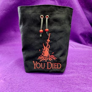 May include: Black dice bag with red drawstring and embroidered image of a bonfire and the text "YOU DIED".
