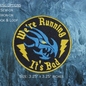 May include: A black and yellow embroidered patch with a blue claw design. The text "We're Running It's Bad" is written in yellow on the patch. The patch is 3.25 inches by 3.25 inches.